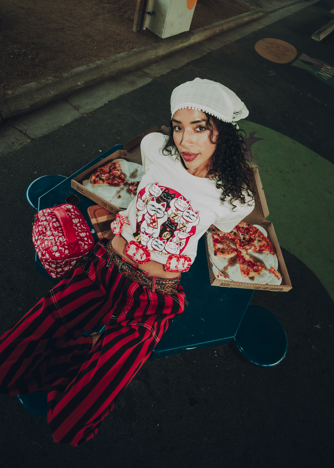 Woman in a white Lucky Cat-themed Pizza John shirt with red pants sitting next to pizzas and a Pizza puffer case