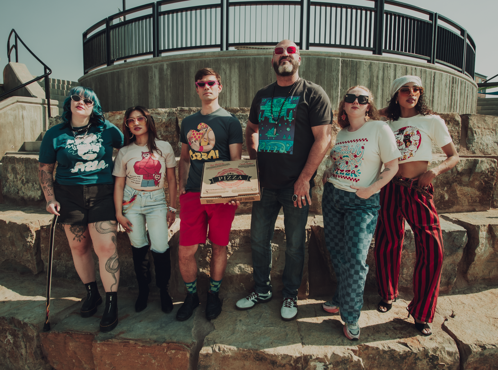 Group of six people posing outdoors on stone steps wearing various Pizzamas shirts