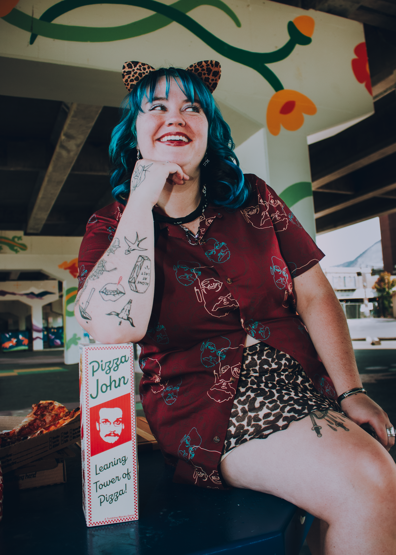 Person with blue hair and tattoos sitting under a colorful mural, wearing a button-up Pizza John shirt, leaning on a Pizza John tumblring blocks box.