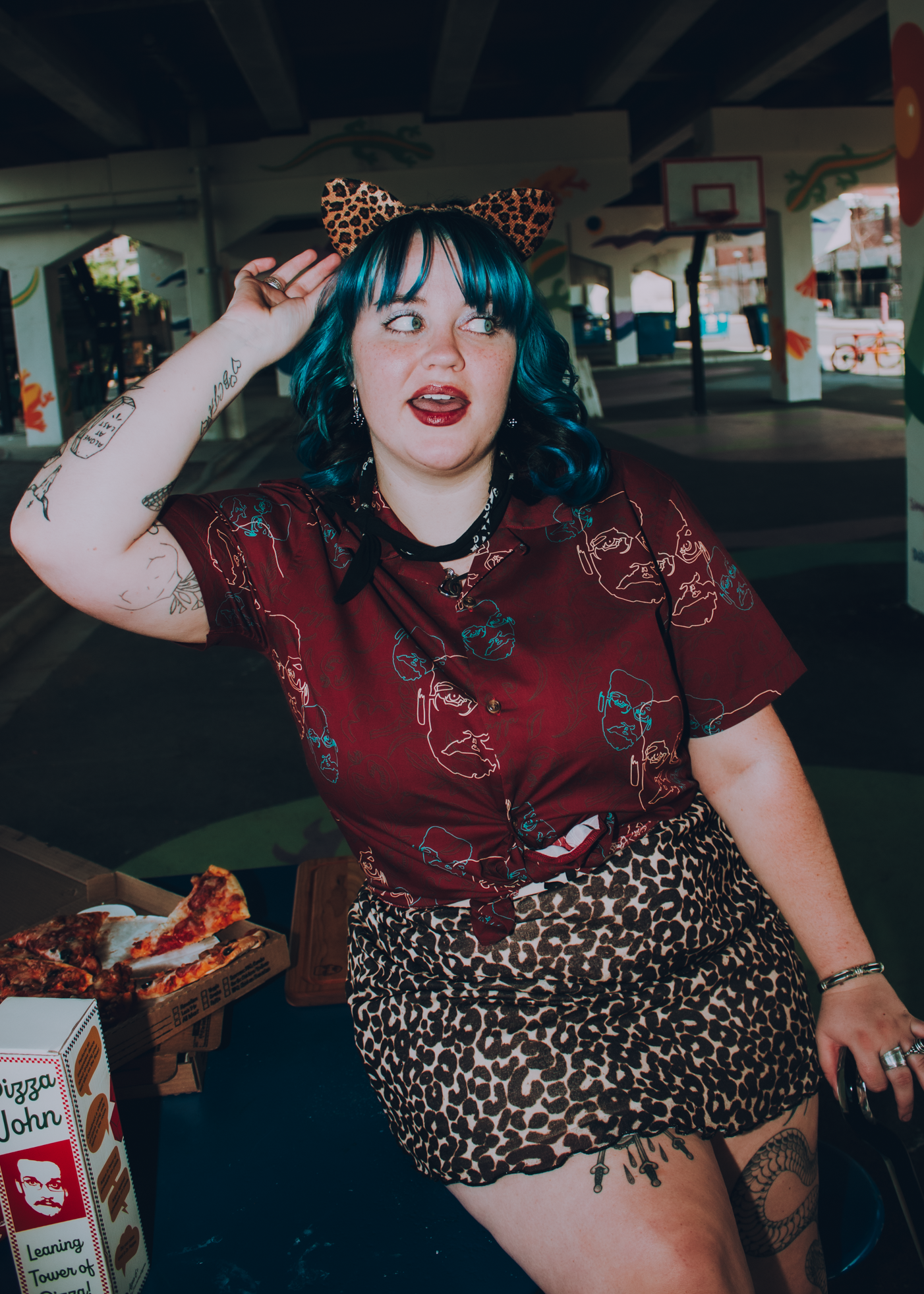 Person with blue hair and leopard print outfit, wearing a Pizza John Button-Up shirt, sitting outdoors near a pizza box.