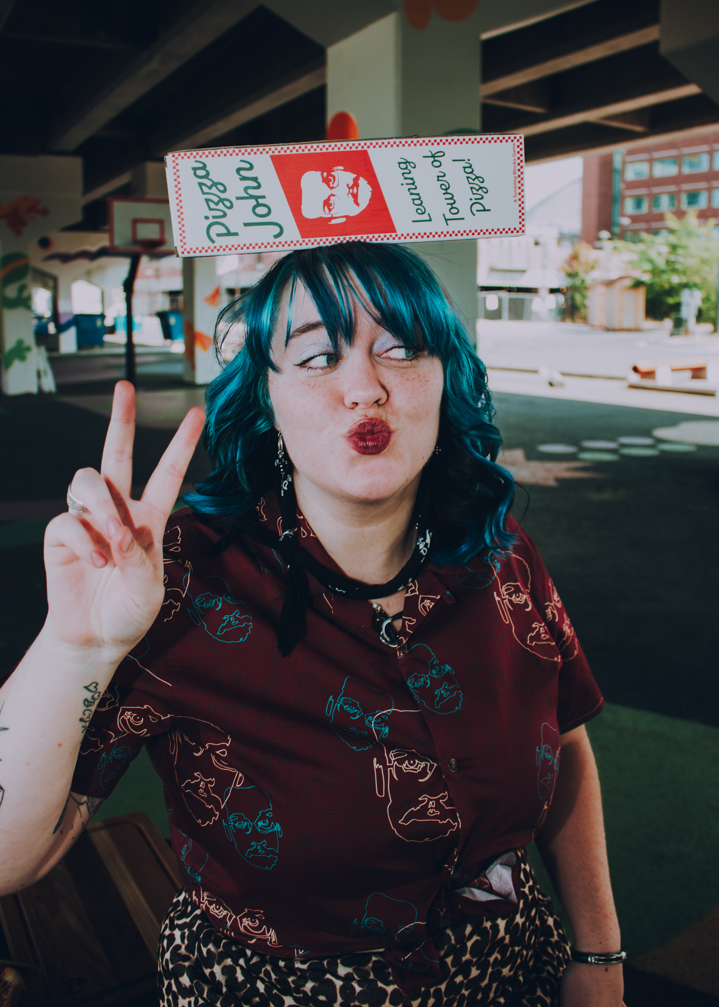 Person with blue hair with a Pizza John Tumbling Block box, wearing a Pizza John button up, and making a peace sign under a bridge.