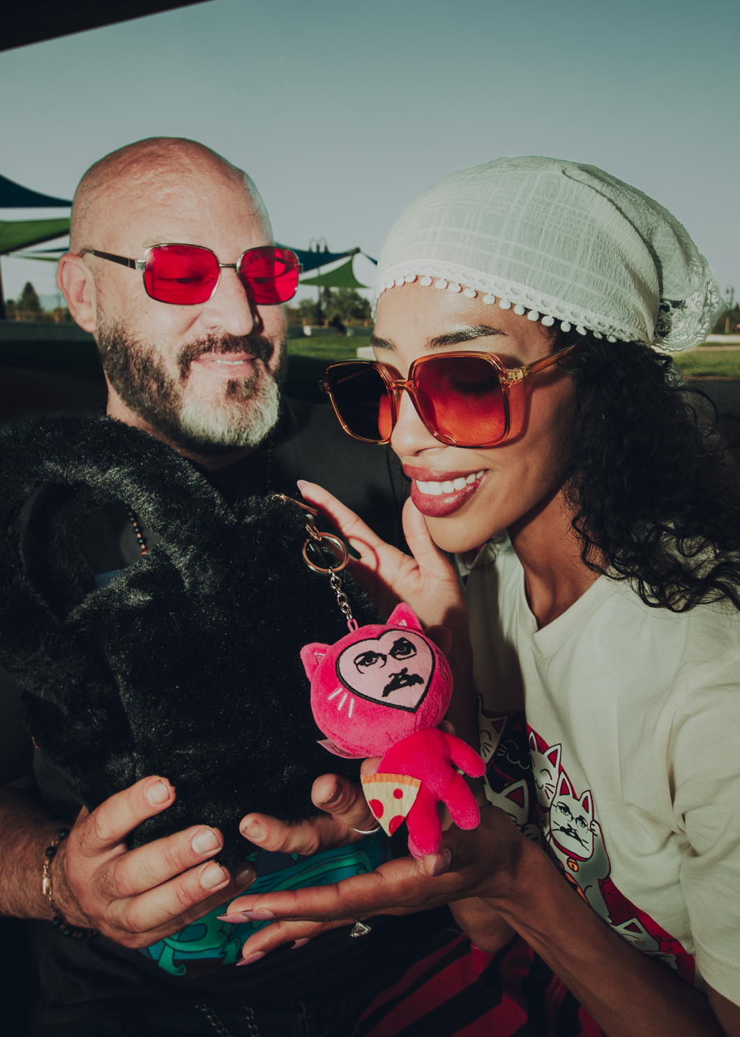 Two people wearing sunglasses, one holding a pink keychain with a pizza john cat design.