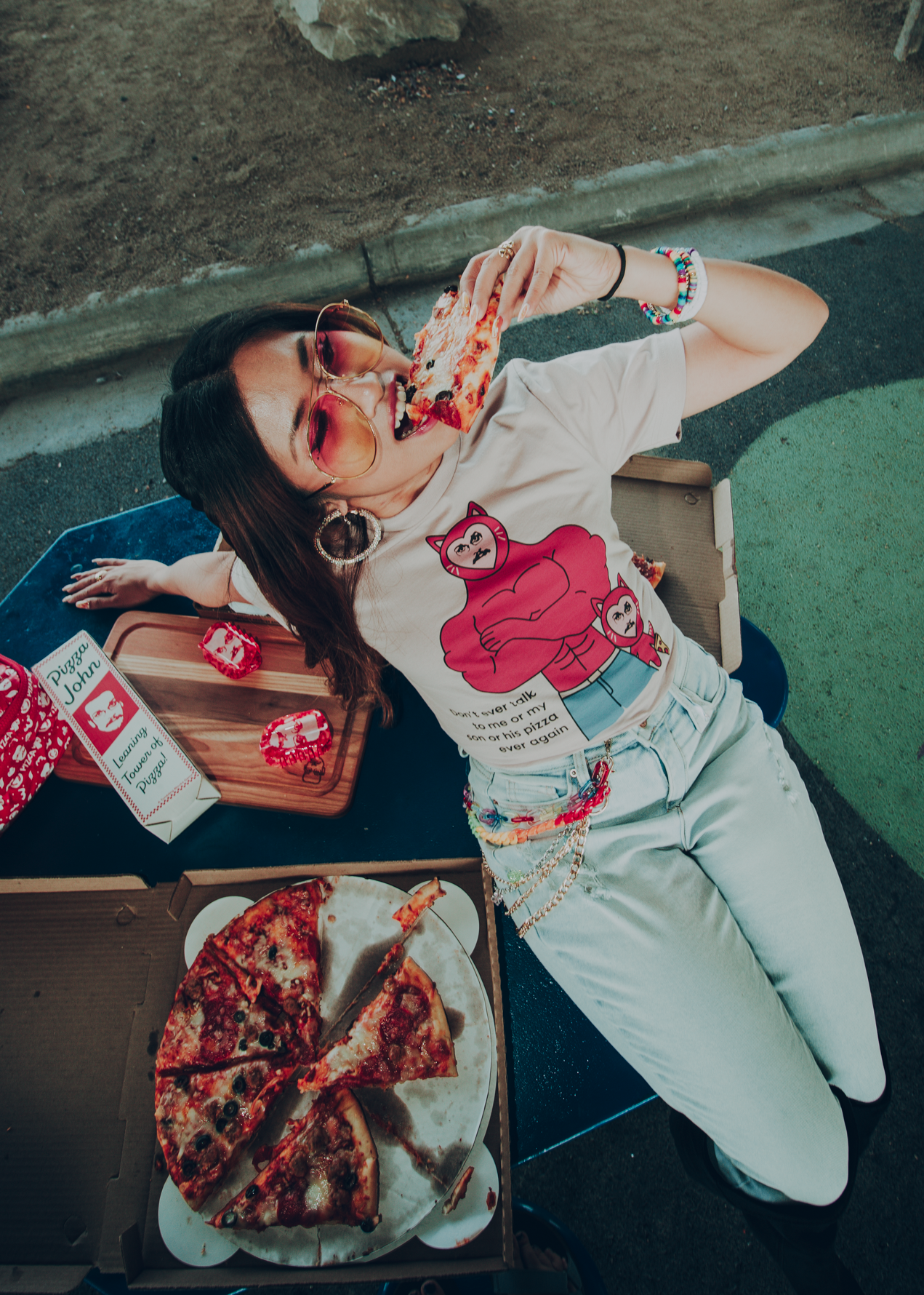 Person eating pizza outdoors wearing a light pink shirt with cartoon cat john figures and text.