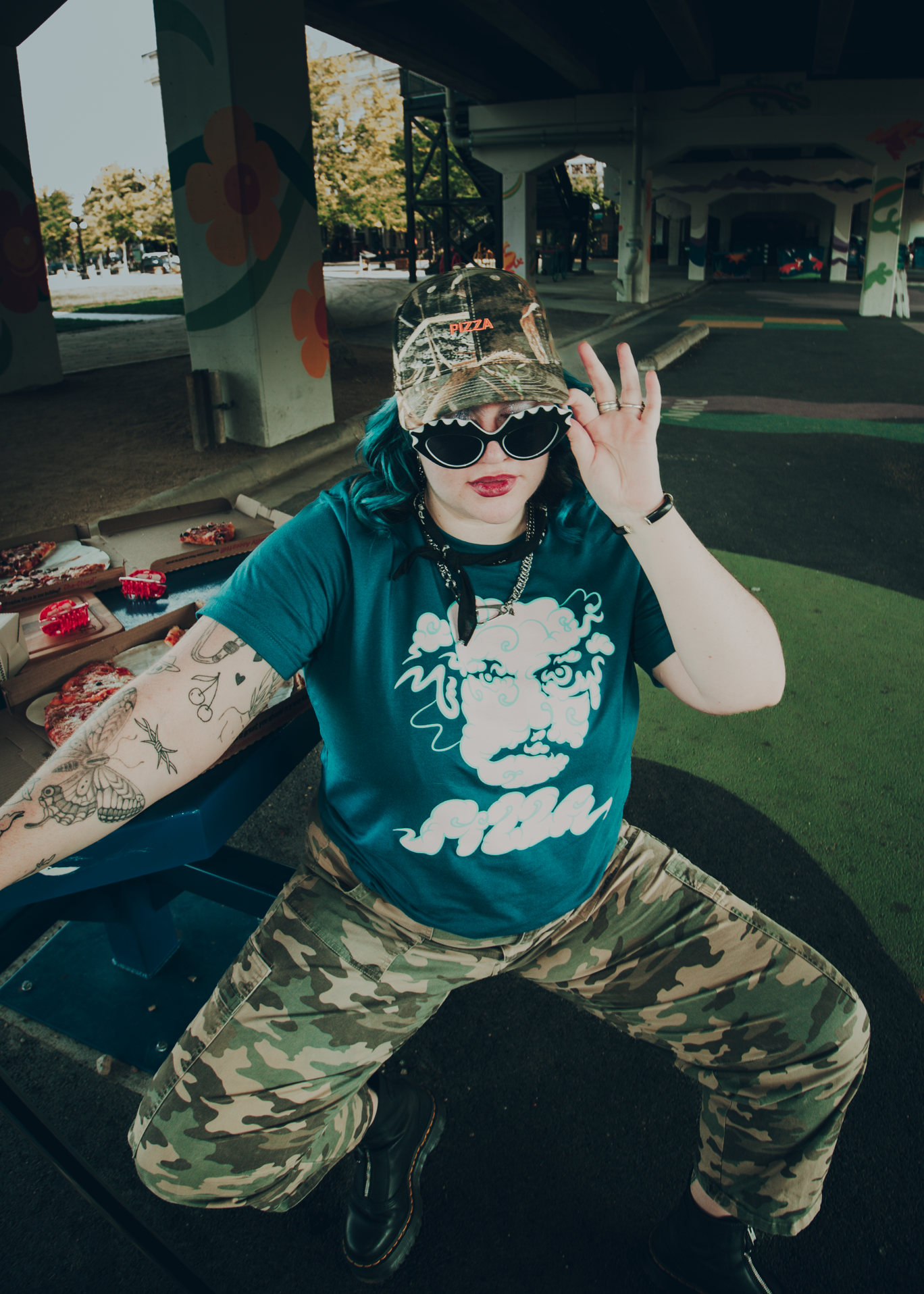 Person wearing a blue t-shirt with a Pizza John face made out of clouds and camouflage pants, sitting on a bench outdoors.