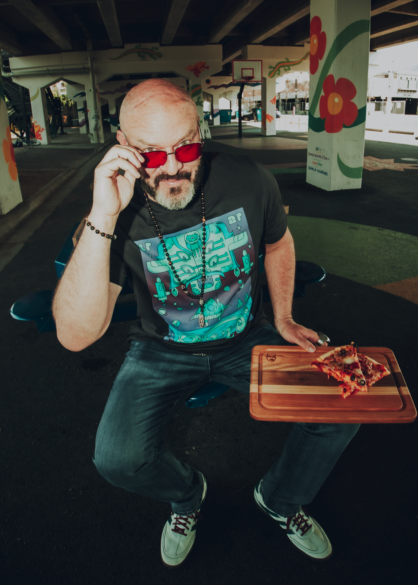 Man in a dark gray video-game themed Pizza John shirtsitting with a pizza on a wooden engraved cutting board in an urban setting