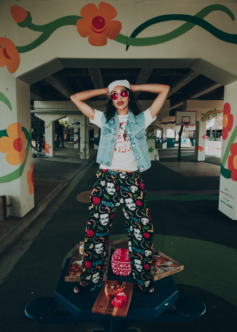 Person posing with arms raised. They are wearing a jean vest with a cat-themed pizza john shirt and vibrant pizzamas-themed pajama pants in a colorful urban setting