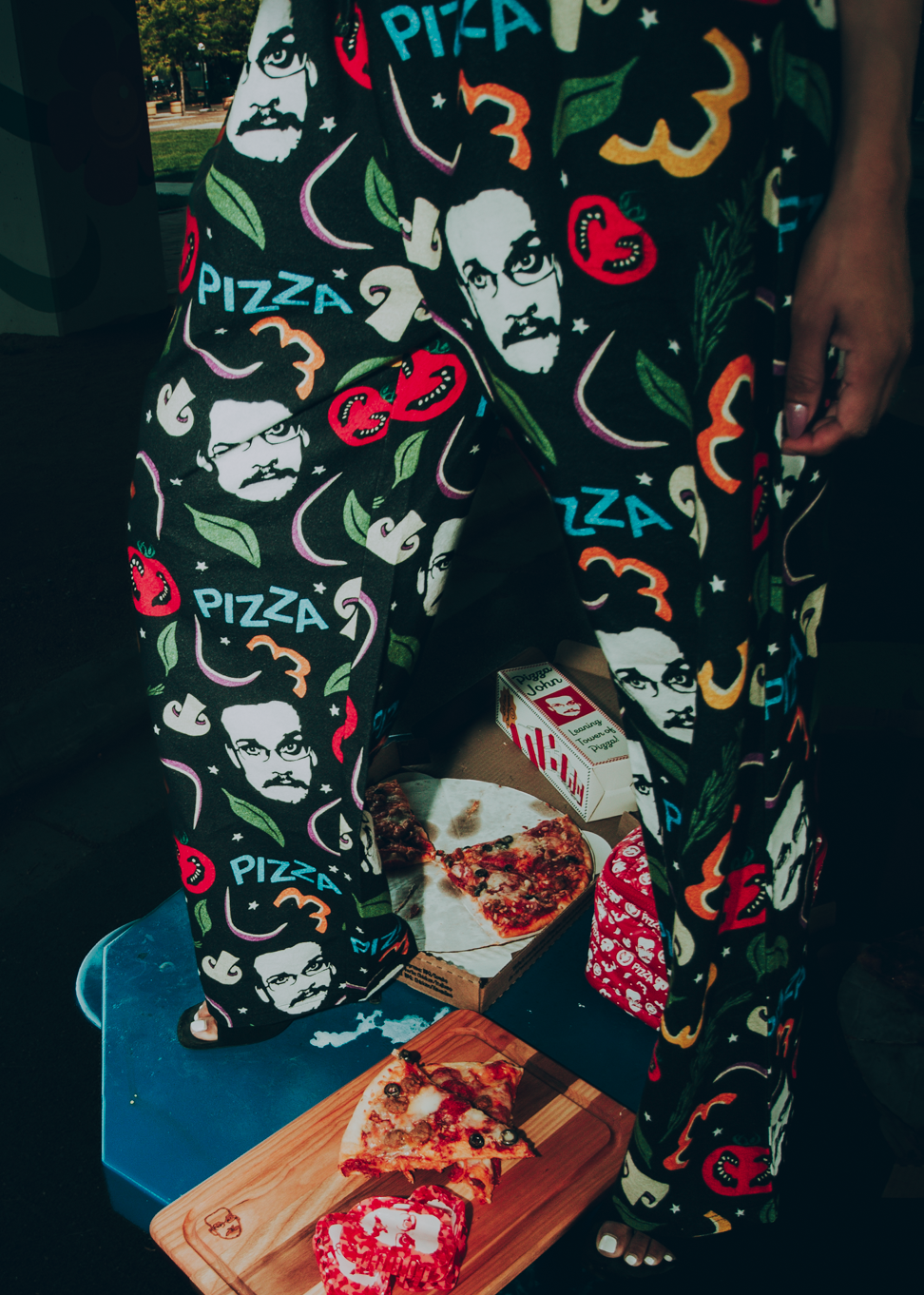 Person wearing a colorful pizza-themed pajama pants, standing over pizza.