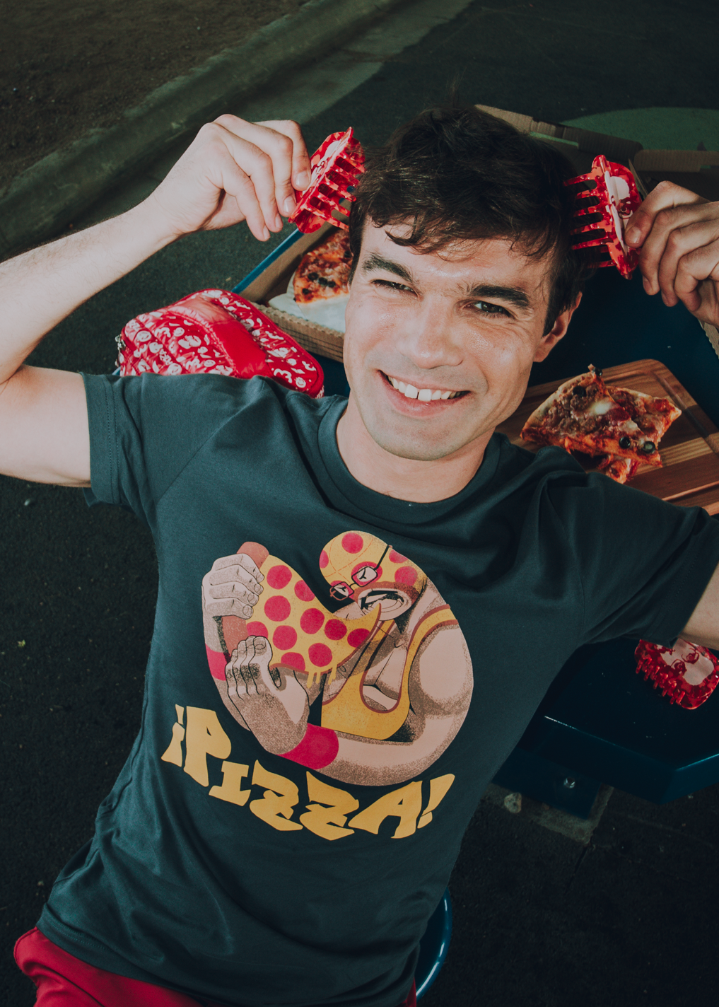 Person wearing a luchador-themed shirt, surrounded by pizzamas products and pizza