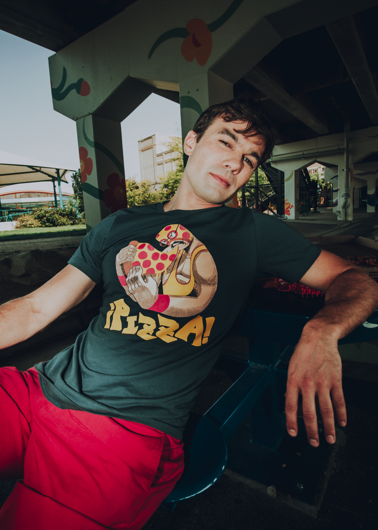 Person wearing a luchador-themed t-shirt with a pizza design sitting outdoors.