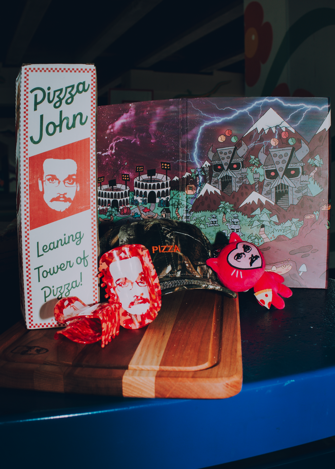 Pizza John items including tumbling blocks, hair claws, camo hat, DM screen, and plushie.
