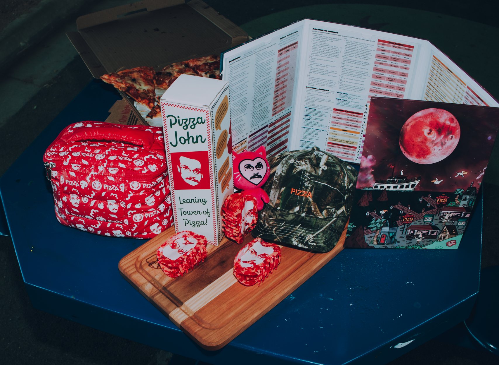 Collection of items including a Pizza John Tumbling Block Game, pizza slices, a plush toy, and a DM screen on a blue surface.