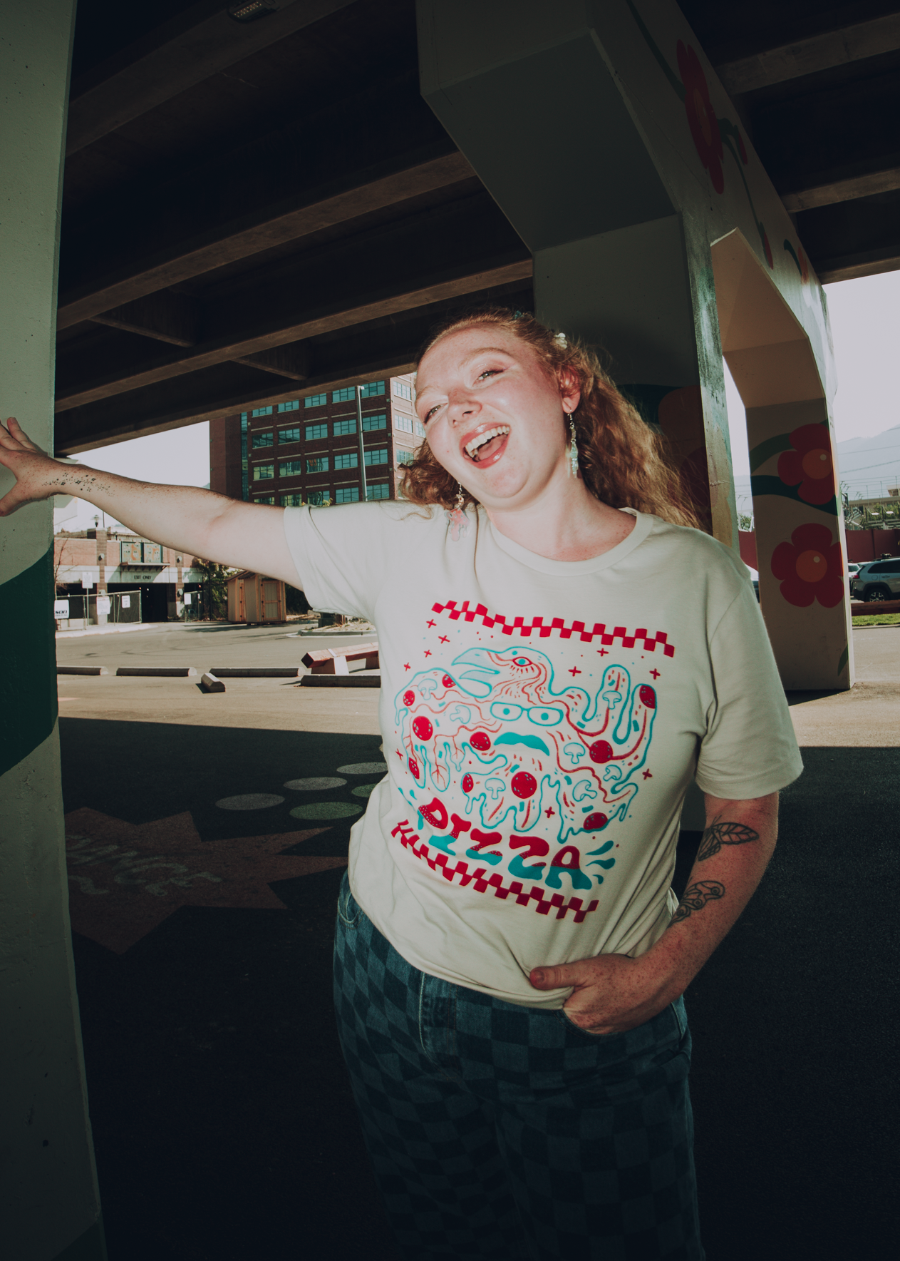 Person wearing a t-shirt with a bird-themed Pizza John design, standing outdoors under a bridge.
