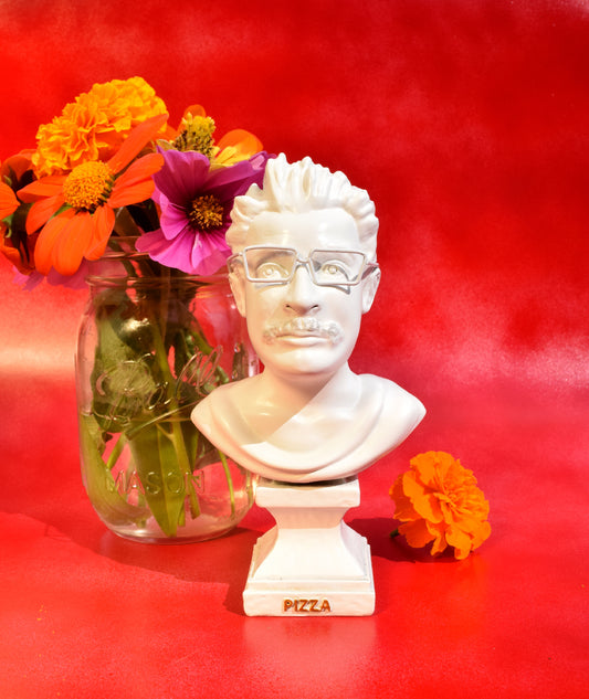 A neoclassical white, mini bust of Pizza John. There is a small square pedestal that has the word pizza on it. The bust features a mustache and glasses. It is sitting in front of a vase of flowers on a red background.