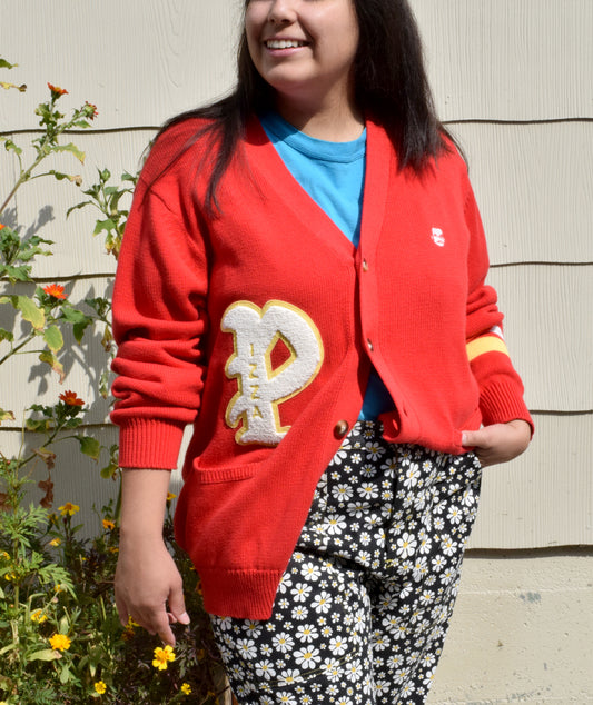 A person wearing a bright red knit cardigan with a big "P" above the right front pocket. There is a small Pizza John head on the left breast pocket area. There is a yellow and white stripe on the the left sleeve. There are buttons up the front.